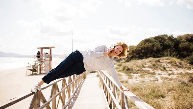 Eine Person in sportlicher Kleidung balanciert seitlich auf einem Holzgeländer einer Strandpromenade und telefoniert dabei. Im Hintergrund sind ein Rettungsturm, das Meer und Dünen zu sehen. Der Himmel ist bewölkt, die Sonne scheint.