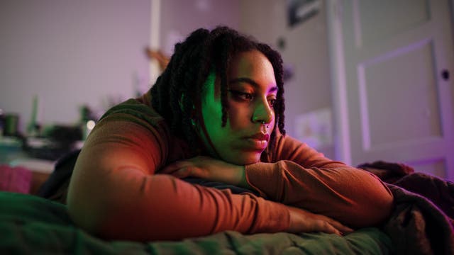 Eine Person liegt nachdenklich auf einem Bett, den Kopf auf die verschränkten Arme gestützt. Der Raum ist in sanftes, farbiges Licht getaucht, das eine ruhige und nachdenkliche Atmosphäre schafft. Im Hintergrund sind unscharfe Details eines Zimmers zu erkennen.