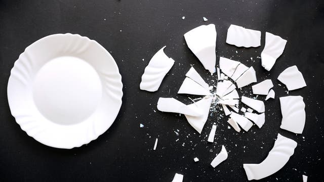 Ein unversehrter weißer Teller liegt auf einer dunklen Oberfläche, während daneben die Scherben eines identischen zerbrochenen Tellers verstreut sind.