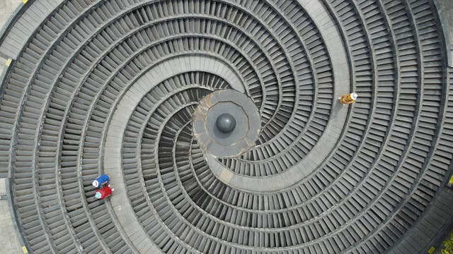 Luftaufnahme einer großen begehbaren Spirale aus grauem Stein, die wie ein Schneckenhaus entworfen wurde. Drei Personen sind von oben erkennbar: Ein Pärchen blau und rot gekleidet im linken Bildteil und eine gelb gekleidete einzelne Person im rechten. Die Szene vermittelt ein Gefühl von Symmetrie und Geometrie.