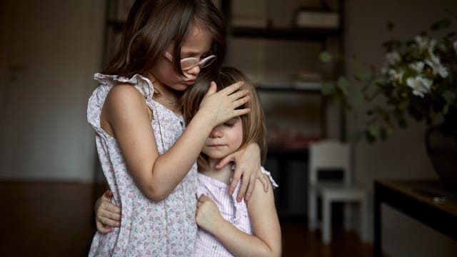 Zwei Kinder in sommerlichen Kleidern stehen in einem Raum. Das ältere Kind umarmt das jüngere und legt tröstend eine Hand auf dessen Kopf. Beide haben die Augen geschlossen. Im Hintergrund sind ein Regal und ein Tisch mit Blumen zu sehen. Die Szene vermittelt ein Gefühl von Geborgenheit und Zuneigung.
