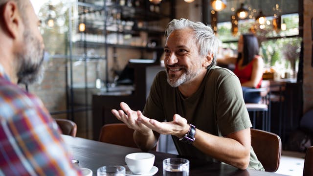 Ein Mann mit grauen Haaren und Bart sitzt in einem Café und unterhält sich lebhaft mit einem anderen Mann, der ihm gegenübersitzt. Auf dem Tisch stehen Kaffeetassen und Gläser mit Wasser. Im Hintergrund ist eine Frau zu sehen, die an einem Tisch sitzt. Die Atmosphäre ist gemütlich und einladend, mit warmem Licht und moderner Einrichtung.