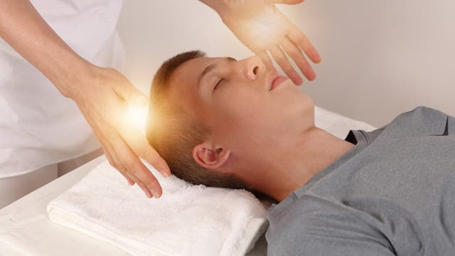 Eine Person liegt entspannt auf einem Tisch mit einem Handtuch unter dem Kopf. Zwei Hände schweben über dem Kopf der Person, umgeben von einem sanften Lichtschein, was auf eine energetische oder therapeutische Behandlung hindeutet. Die Szene vermittelt Ruhe und Entspannung.