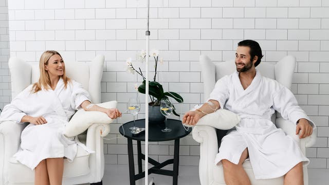 Zwei Personen in weißen Bademänteln sitzen in bequemen Sesseln in einem hellen Raum mit weißen Fliesenwänden. Beide erhalten eine Infusion über einen intravenösen Zugang am Arm. Zwischen ihnen steht ein kleiner Tisch mit einer Orchidee und zwei Gläsern Wasser. Sie lächeln und wirken entspannt.