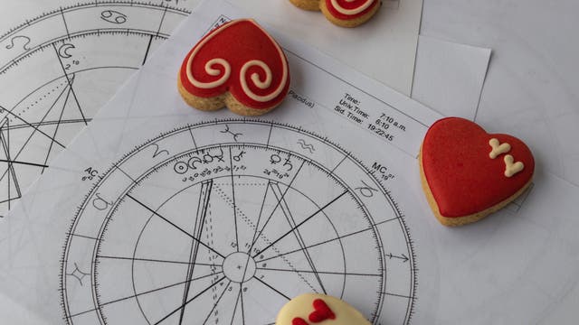 Ein Bild zeigt astrologische Diagramme mit verschiedenen Symbolen und Linien, die auf Papier gedruckt sind. Auf den Diagrammen liegen herzförmige Kekse mit rotem Zuckerguss und dekorativen Mustern. Die Kekse sind in verschiedenen Designs gestaltet, darunter Spiralen und kleine Herzen. Das Bild kombiniert wissenschaftliche und dekorative Elemente und vermittelt eine Mischung aus Astrologie und kulinarischer Kunst.
