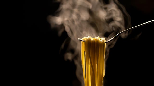 Eine Gabel hält dampfende Spaghetti vor einem dunklen Hintergrund. Der Dampf steigt in weichen Schwaden auf, was auf die Frische und Wärme der Pasta hinweist. Die Beleuchtung hebt die goldene Farbe der Spaghetti hervor.