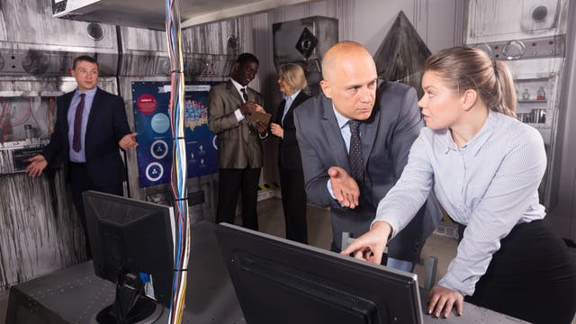 Eine Gruppe von Personen in einem Raum, der wie ein Kontrollzentrum aussieht. Zwei Personen im Vordergrund diskutieren angeregt über einen Computerbildschirm. Im Hintergrund sind weitere Personen zu sehen, die miteinander sprechen und Notizen machen. An den Wänden befinden sich technische Geräte und ein Poster mit Diagrammen. Die Szene vermittelt eine geschäftige und konzentrierte Arbeitsatmosphäre.