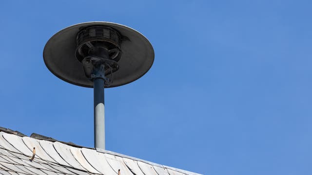 Ein grauer Sirenenmast ragt über ein mit Schieferplatten gedecktes Dach in den blauen Himmel. Die Sirene hat eine runde, schalenförmige Abdeckung. Das Bild zeigt eine klare, wolkenlose Wetterlage.