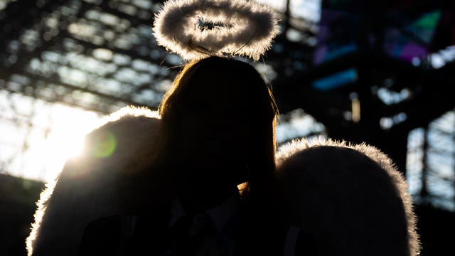 Eine Person in einem Engelskostüm mit Heiligenschein und Flügeln steht im Gegenlicht. Die schwarze Silhouette hebt sich vor einer strukturierten, lichtdurchfluteten Glasdecke ab. Die Szene vermittelt eine mystische Atmosphäre durch das Spiel von Licht und Schatten.