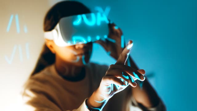 Eine Person trägt ein Virtual-Reality-Headset und interagiert mit einer digitalen Benutzeroberfläche. Der Hintergrund ist in Blau- und Orangetönen gehalten, und leuchtende Zahlen oder Buchstaben sind sichtbar. Die Person zeigt mit dem Finger auf etwas in der virtuellen Umgebung, was auf eine immersive Erfahrung hinweist.