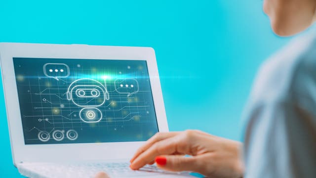 Eine Person arbeitet an einem Laptop mit einem blauen Bildschirm, auf dem ein digitales Symbol eines Chatbots mit Sprechblasen und Schaltkreisen angezeigt wird. Die Person tippt auf der Tastatur. Das Bild vermittelt das Thema künstliche Intelligenz und digitale Kommunikation.