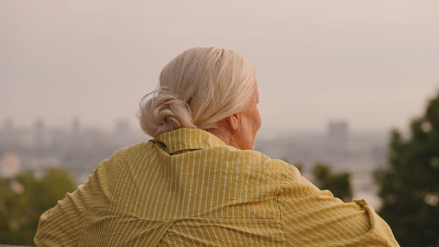 Eine ältere Person mit weißem Haar und einem Dutt steht mit dem Rücken zur Kamera und blickt auf eine verschwommene Stadtlandschaft in der Ferne. Im Hintergrund sind Bäume und Gebäude zu erkennen.