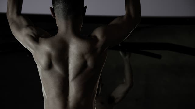 Eine Person mit muskulösem Rücken macht Klimmzüge an einer Stange in einem Fitnessstudio. Die Szene ist dunkel beleuchtet, wodurch die Definition der Rückenmuskulatur hervorgehoben wird. Es sind keine weiteren Details oder Texte im Bild vorhanden.
