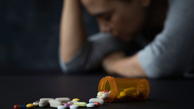 Eine unscharf zu sehende Frau sitzt mit gesenktem Kopf im Hintergrund, während im Vordergrund ein umgefallenes Pillenfläschchen mit verstreuten Tabletten zu sehen ist. Die Szene vermittelt ein Gefühl von Sorge oder Verzweiflung.