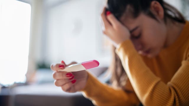 Eine Frau sitzt auf einem Sofa und hält einen Schwangerschaftstest in der Hand. Sie stützt ihren Kopf mit der anderen Hand und wirkt enttäuscht. 