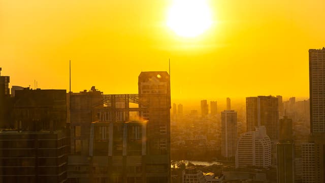 Städtische Skyline bei Sonnenuntergang, mit hohen Gebäuden im Vordergrund und einer leuchtend gelben Sonne am Himmel. Die Szene ist in warmes, goldenes Licht getaucht, das die Silhouetten der Wolkenkratzer betont.