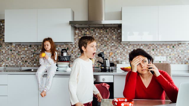 Eine Frau sitzt in einer Küche und telefoniert, während sie sich die Stirn hält, was auf Stress oder Erschöpfung hindeutet. Ein Kind steht neben ihr und scheint zu rufen oder zu singen. Ein weiteres Kind sitzt auf der Arbeitsplatte und hält eine Orange. Die Küche ist modern eingerichtet mit weißen Schränken und einer Mosaikfliesenrückwand. Auf dem Tisch steht eine rote Tasse mit weißen Punkten neben einem Laptop.