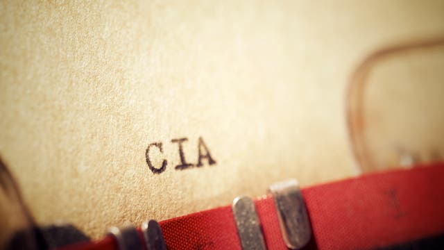 Eine Nahaufnahme einer Schreibmaschine, die das Wort "CIA" auf ein beiges Blatt Papier tippt. Der Fokus liegt auf den getippten Buchstaben, während der Rest des Bildes leicht verschwommen ist. Die Szene vermittelt ein Gefühl von Geheimhaltung und klassischer Büroarbeit.