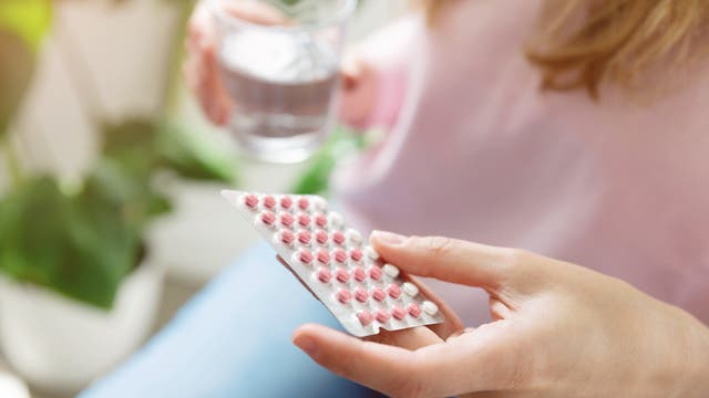 Eine Frau hält eine Blisterpackung mit vier Reihen rosa und einer Reihe weißen Tabletten in der einen Hand und ein Glas Wasser in der anderen. Im Hintergrund sind unscharf grüne Pflanzen zu sehen. Die Szene vermittelt den Eindruck der Einnahme von Medikamenten.