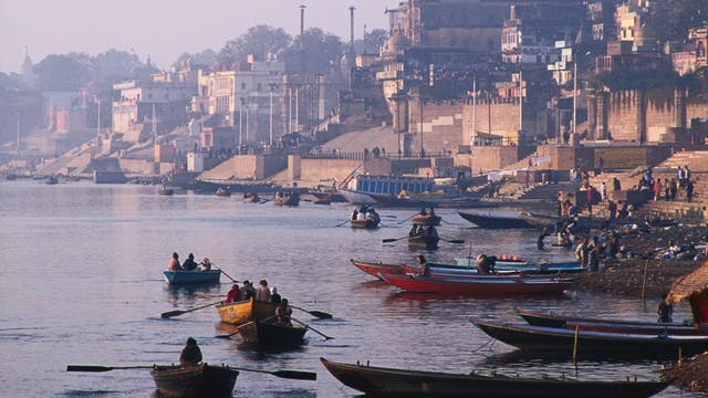 Eine belebte Flusslandschaft mit mehreren Booten, die auf dem Wasser treiben. Im Hintergrund sind historische Gebäude und Treppen zu sehen, die zum Fluss hinabführen. Menschen sind auf den Booten und entlang des Ufers zu erkennen, einige scheinen zu rudern, während andere am Ufer verweilen. Die Szene vermittelt eine geschäftige und kulturell reiche Atmosphäre.
