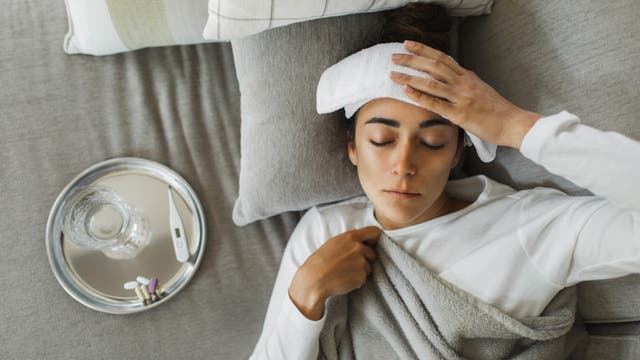 Eine Person liegt auf einem grauen Sofa und hat ein feuchtes Tuch auf der Stirn. Neben ihr steht ein Tablett mit einem Glas Wasser, einem Thermometer und Medikamenten. Die Person ist in eine Decke gehüllt und scheint sich auszuruhen. Die Szene vermittelt den Eindruck von Ruhe und Genesung.