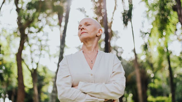 Eine Frau mit einer Glatze steht in einem Wald und hat die Augen geschlossen.