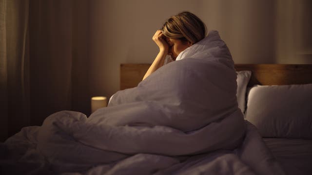 Eine junge Frau sitzt in eine Decke gewickelt im Bett und hält sich den Kopf