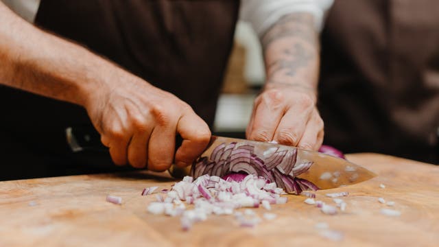 Eine Person schneidet eine rote Zwiebel auf einem Holzbrett mit einem großen Küchenmesser. Die Zwiebel ist in feine Würfel geschnitten. Die Person trägt eine braune Schürze. Im Hintergrund ist eine weitere Person unscharf zu sehen.