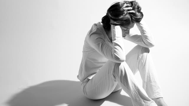 Eine Person sitzt auf dem Boden in einem hellen Raum, die Knie angezogen und den Kopf in den Händen vergraben. Die Person trägt helle Kleidung, und der Schatten fällt deutlich auf den Boden. Die Szene vermittelt einen Ausdruck von Nachdenklichkeit oder Stress.