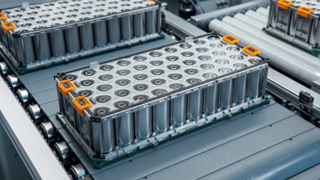 Eine Nahaufnahme eines Batteriepakets auf einem Förderband in einer Fabrik. Die Batterien sind in einem rechteckigen Metallgehäuse angeordnet, mit orangefarbenen Griffen an den Ecken. Das Bild zeigt den industriellen Herstellungsprozess von Batterien, möglicherweise für Elektrofahrzeuge oder Energiespeicherlösungen. Keine Menschen sind im Bild zu sehen.
