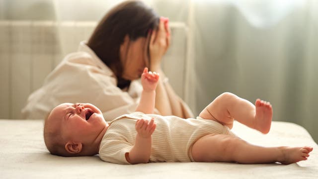 Ein Baby liegt auf einem Bett und weint. Im Hintergrund sieht man die verzweifelte Mutter am Boden sitzen.