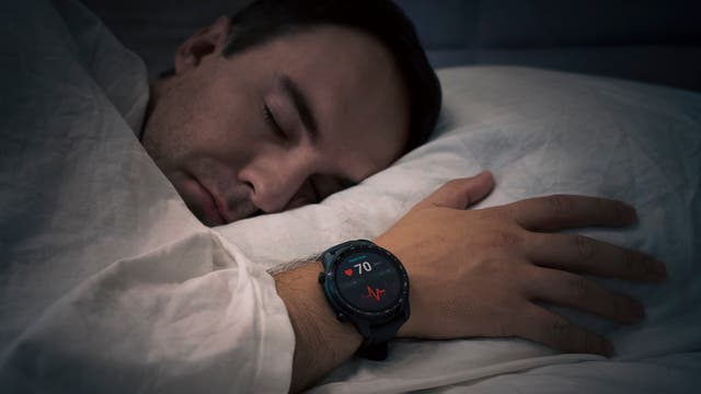 Ein Mann mit einer Smartwatch am Handgelenk liegt im Bett und tut so, als würde er schlafen.