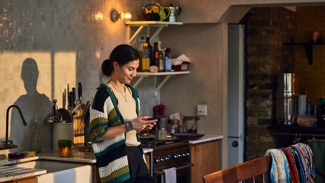 Eine Person steht in einer gemütlichen Küche und schaut auf ihr Smartphone. Die Küche ist mit einem Herd, Regalen mit Gewürzen und einer Spüle ausgestattet. Sonnenlicht fällt durch ein Fenster und wirft Schatten an die Wand. Die Atmosphäre wirkt warm und einladend.