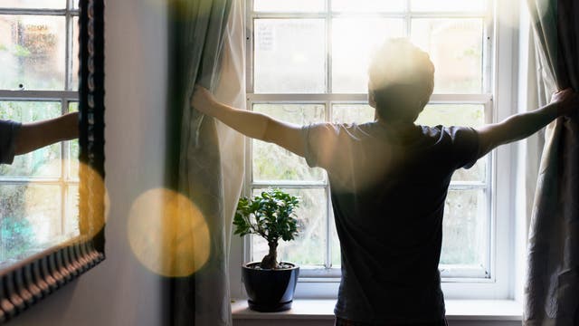 Eine Person steht mit dem Rücken zur Kamera und zieht die Vorhänge eines großen Fensters auf, durch das Sonnenlicht hereinfällt. Auf der Fensterbank steht eine kleine Topfpflanze. Die Szene wirkt ruhig und einladend, mit einem Fokus auf Morgenlicht und Natur.
