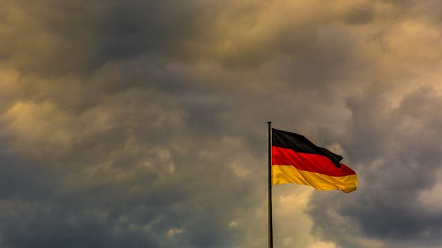Eine deutsche Flagge weht im Wind vor einem dramatischen, wolkenverhangenen Himmel. Die Farben Schwarz, Rot und Gold sind deutlich sichtbar. Die Szene vermittelt eine Stimmung von Stärke und Beständigkeit.