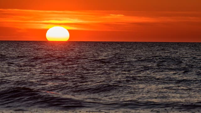 Sonnenuntergang über dem Meer mit einem leuchtend orangefarbenen Himmel. Die Sonne steht tief am Horizont und spiegelt sich im welligen Wasser. Keine Menschen oder Text im Bild.