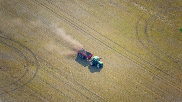 Ein grüner Traktor zieht einen roten Anhänger über ein weitläufiges Feld und hinterlässt eine Staubwolke. Das Bild zeigt landwirtschaftliche Aktivitäten aus der Vogelperspektive, wobei die gleichmäßigen Linien des Feldes sichtbar sind. Es gibt keine Menschen im Bild.