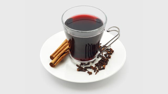 Ein Glas mit rotem Glühwein, umgeben von Zimtstangen und Nelken, auf einem grauen Hintergrund. Das Glas hat einen Metallgriff. 