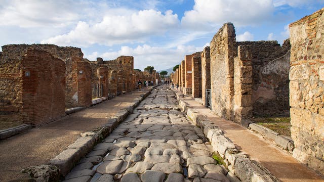 Eine antike, gepflasterte Straße in Pompeji, gesäumt von Ruinen aus Stein und Ziegeln. Der Himmel ist teils bewölkt, und einige Menschen sind in der Ferne zu sehen, die die historische Stätte erkunden. Die Szene vermittelt ein Gefühl von Geschichte und archäologischer Bedeutung.