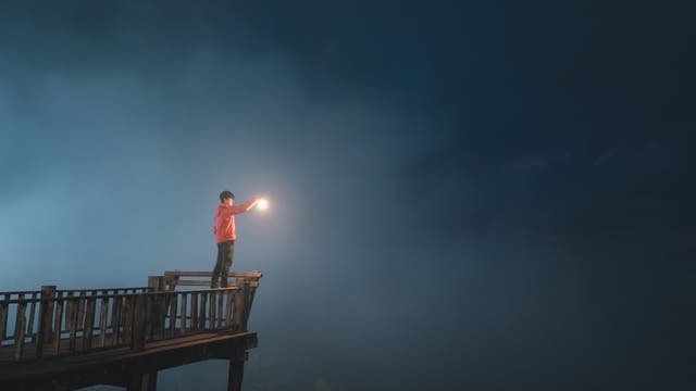 Eine Person steht auf einem Holzsteg und hält eine leuchtende Laterne in der Hand. Der Hintergrund ist dunkel und neblig, was eine mystische Atmosphäre erzeugt. Die Person trägt eine rote Jacke und blickt in die Ferne.