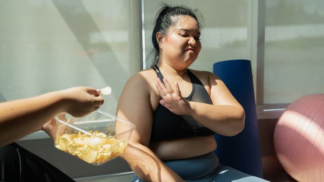 Eine Person in Sportkleidung sitzt in einem Raum mit Fitnessgeräten und lehnt eine Schale mit Chips ab, die ihr von einer anderen Person angeboten wird. Die Szene deutet auf eine gesunde Lebensweise oder eine bewusste Ernährungsentscheidung hin. Im Hintergrund sind eine Yogamatte und ein Gymnastikball zu sehen.