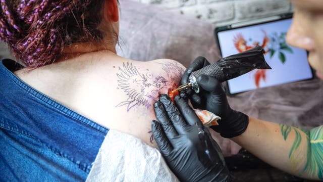 Eine Person erhält ein Tattoo auf der Schulter, das einen Vogel mit ausgebreiteten Flügeln zeigt. Der Tätowierer trägt schwarze Handschuhe und verwendet eine Tätowiermaschine. Im Hintergrund ist ein Tablet mit einer farbigen Darstellung des Vogels zu sehen. Die Szene findet in einem Studio mit einer weißen Backsteinwand statt.