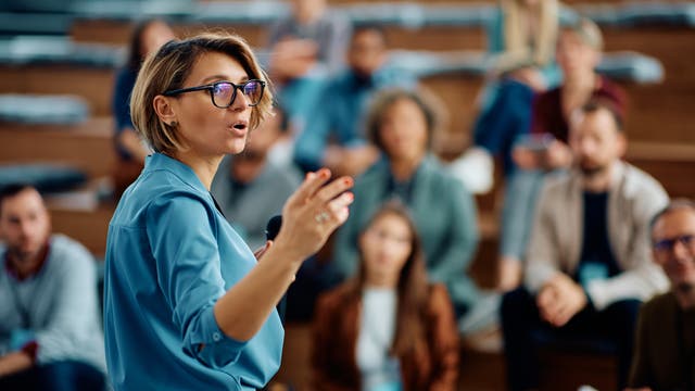 Eine Hochschullehrerin mit Mikrofon steht vor Studierenden in einem Hörsaal