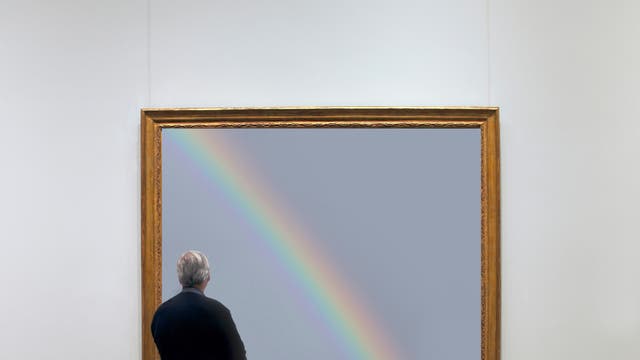 Eine Person steht vor einem großen Gemälde in einem Museum. Das Gemälde zeigt einen Regenbogen auf grauem Hintergrund und ist in einem goldenen Rahmen eingefasst. Die Person betrachtet das Kunstwerk aufmerksam.