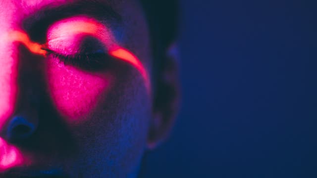 Nahaufnahme eines Gesichts mit geschlossenen Augen, beleuchtet von intensivem pinkem und blauem Licht. Ein roter Lichtstreifen verläuft diagonal über das Auge. Die Szene wirkt künstlerisch und futuristisch. Keine Text- oder URL-Informationen vorhanden.
