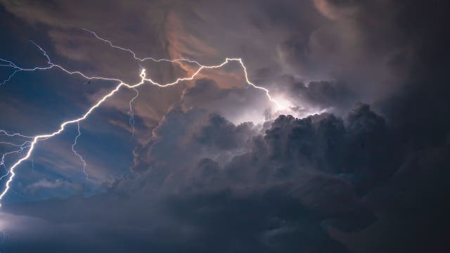Ein nächtlicher Himmel mit dramatischen Gewitterwolken, durchzogen von mehreren hellen Blitzen. Die Blitze erhellen die dunklen Wolken und erzeugen ein beeindruckendes Naturschauspiel. Am unteren Rand des Bildes sind Silhouetten von Bäumen zu sehen.