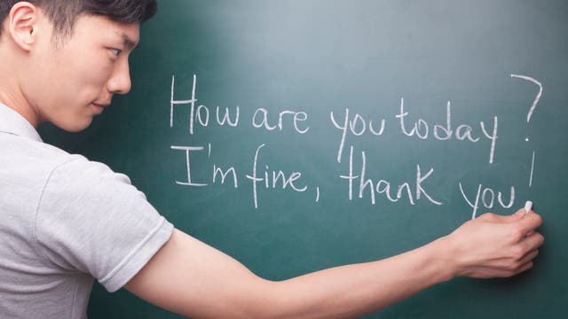 Ein Mann schreibt mit Kreide auf eine grüne Tafel. Der Text auf der Tafel lautet: "How are you today? I'm fine, thank you!" Der Mann trägt ein helles T-Shirt und konzentriert sich auf das Schreiben.