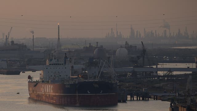 Ein großes Frachtschiff liegt in einem Industriehafen vor Anker. Im Hintergrund sind zahlreiche Fabrikanlagen und Schornsteine zu sehen, aus denen Rauch aufsteigt. Die Szene ist in ein gedämpftes Licht getaucht, das eine dunstige Atmosphäre erzeugt. Stromleitungen verlaufen über den Hafen, und Kräne sind entlang der Uferlinie sichtbar.