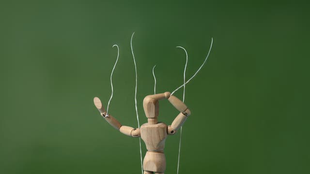 Eine hölzerne Gliederpuppe, die an Fäden aufgehängt ist, steht vor einem blauen Hintergrund. Die Puppe hebt einen Arm, während der andere Arm an der Seite bleibt. Die Fäden sind an Kopf, Armen und Beinen befestigt, was den Eindruck einer Marionette vermittelt.