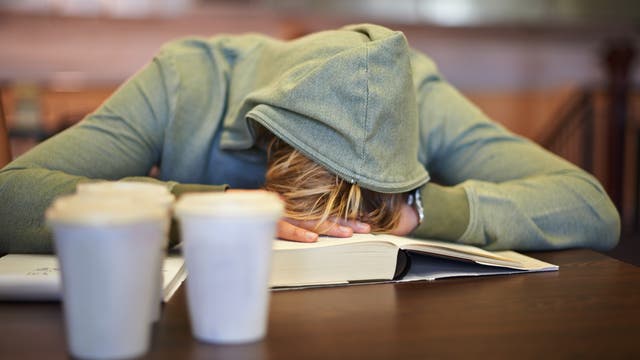 Eine Person in einem grünen Hoodie mit aufgezogener Kapuze sitzt an einem Tisch und hat den Kopf auf ein aufgeschlagenes Buch gelegt, als ob sie schläft oder erschöpft ist. Im Vordergrund stehen zwei Kaffeebecher.